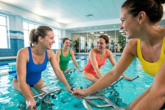 Groupe de femmes adultes pratiquant l'aquabiking ensemble dans une piscine intérieure moderne, souriant et pédalant en rythme sur des vélos aquatiques colorés